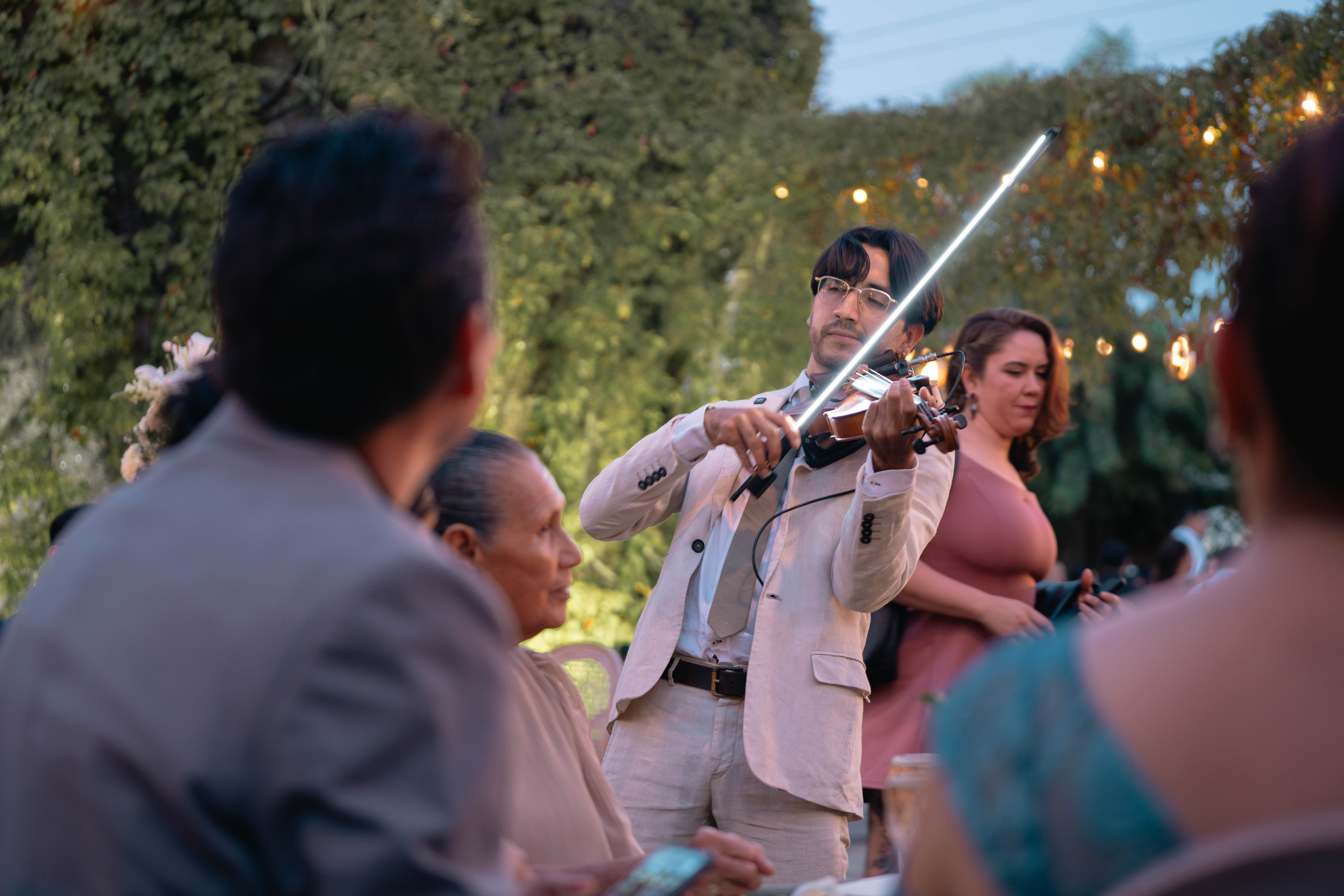 Yael Magaña tocando violín con arco luminoso en una boda, con invitados alrededor